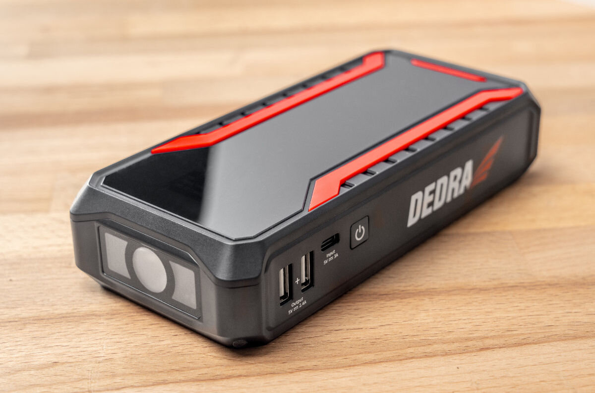Na drewnianym blacie leży czarny powerbank z czerwonymi akcentami i napisem \'DEDRA\'. Urządzenie posiada dwa porty USB, port micro USB oraz przycisk zasilania.