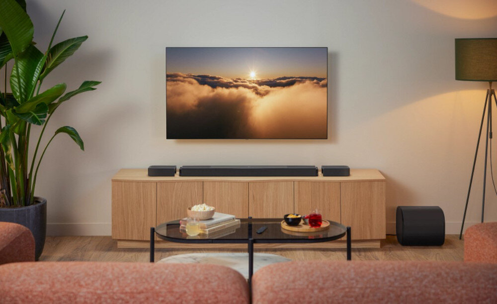 W salonie znajduje się telewizor wyświetlający obraz chmur o zachodzie słońca, umieszczony na ścianie nad drewnianą szafką. Przed szafką stoi soundbar, a obok niego rośliny doniczkowe i lampa stojąca.