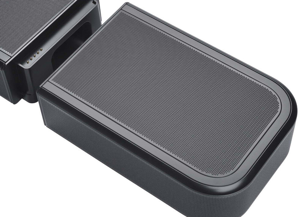 Na obrazie widoczny jest czarny soundbar z metalową siatką na górnej powierzchni. Obok znajduje się drugi element o podobnym wyglądzie, z widocznymi złączami na boku.