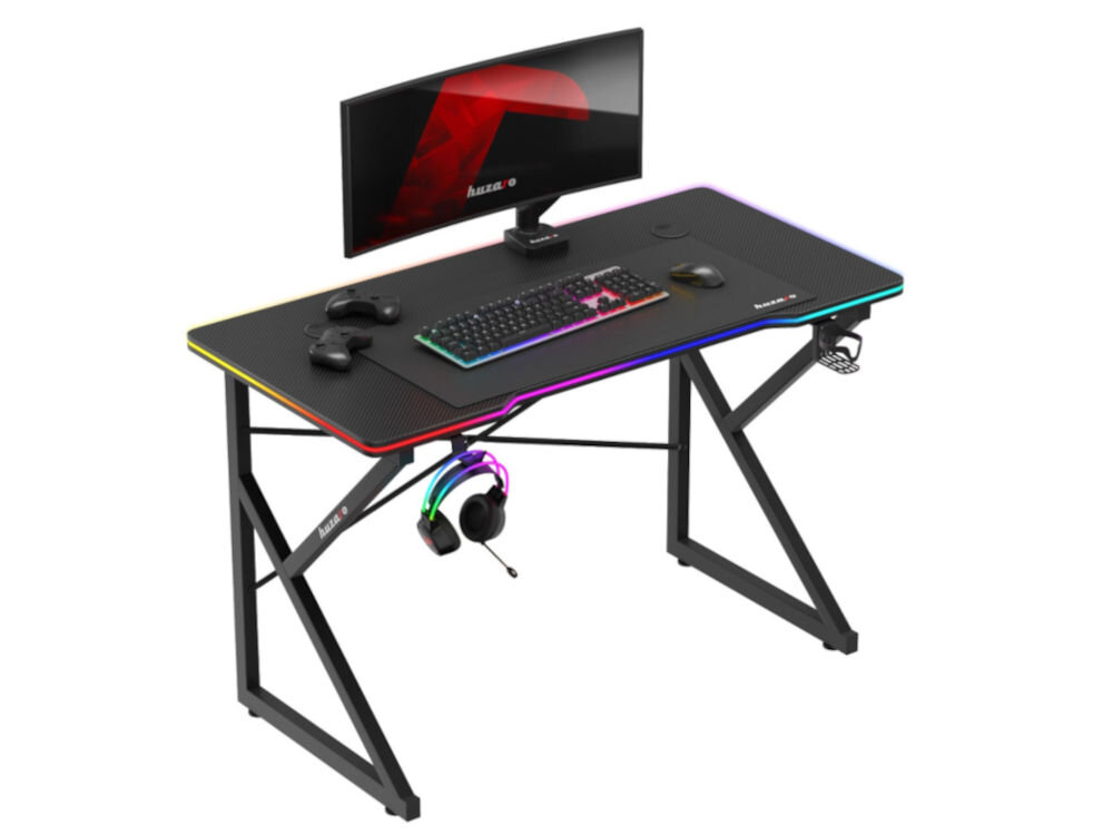 Czarne biurko gamingowe z kolorowym podświetleniem krawędzi, na którym znajduje się monitor, klawiatura, myszka i kontrolery. Na biurku widoczne jest logo \'huzaro\'.