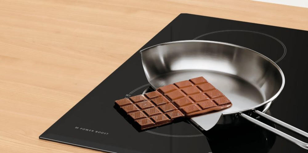 Na czarnej płycie kuchennej znajduje się metalowa patelnia z tabliczką czekolady. Na płycie widoczny jest napis \'HI POWER BOOST\'.