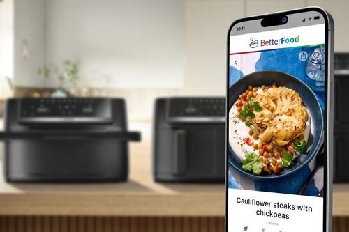 Na pierwszym planie widoczny jest smartfon z aplikacją BetterFood, pokazującą danie z kalafiora i ciecierzycy. W tle znajdują się dwa czarne urządzenia kuchenne, prawdopodobnie frytkownice beztłuszczowe.