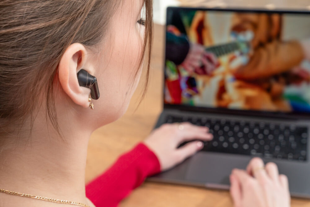 Osoba nosi czarne słuchawki douszne, patrząc na ekran laptopa, na którym wyświetlany jest obraz z gitarą. Osoba ma na sobie czerwoną bluzkę.