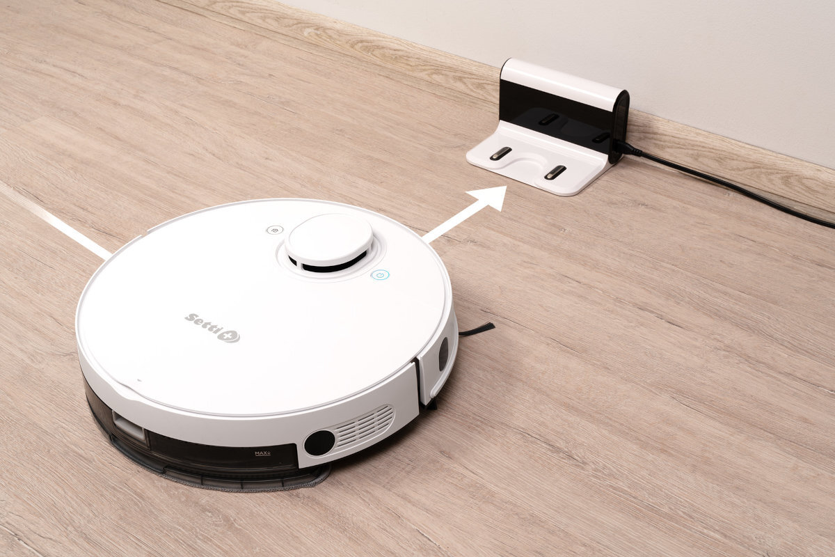 Na drewnianej podłodze znajduje się biały robot sprzątający z napisem \'Setti+\'. W tle widoczna jest stacja dokująca podłączona do gniazdka elektrycznego.