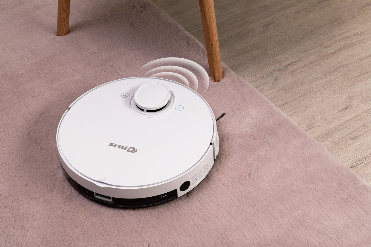 Na obrazie widoczny jest biały robot sprzątający z logo \'Setti\' na dywanie. Obok robota znajduje się drewniana noga mebla na podłodze.