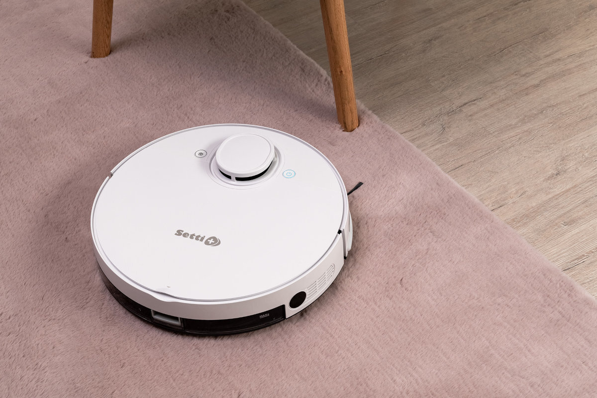 Na obrazie widoczny jest biały robot sprzątający z logo \'Setti\' na górze, poruszający się po różowym dywanie obok drewnianej nogi mebla.