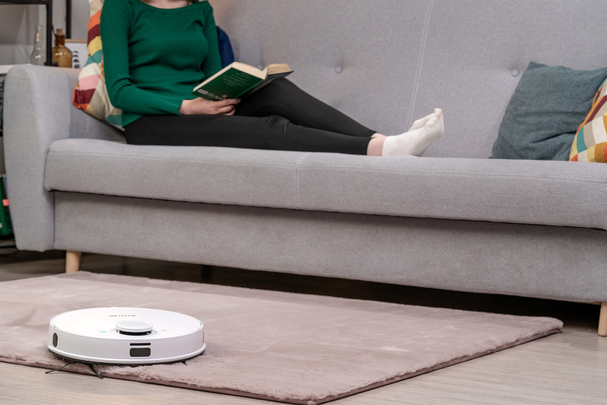 Na dywanie znajduje się biały robot sprzątający. W tle widać osobę siedzącą na kanapie z książką w ręku.