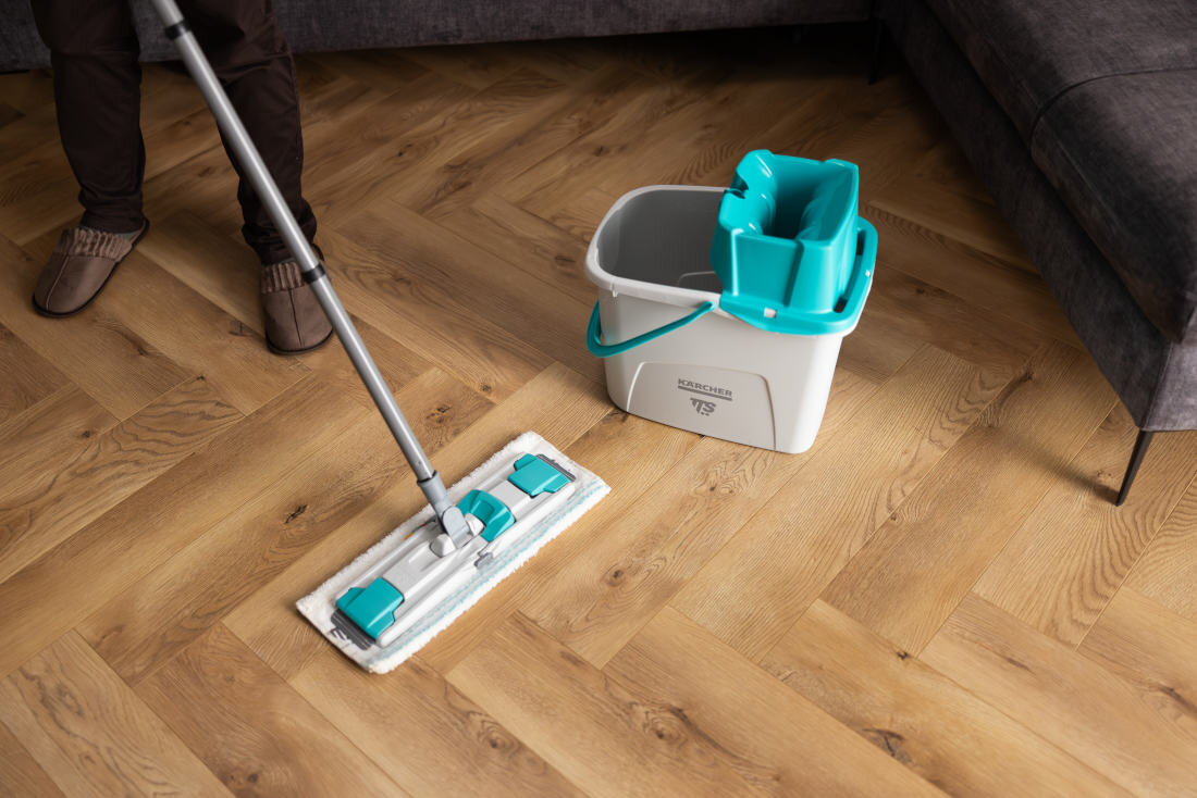 Osoba używa mopa płaskiego na drewnianej podłodze, obok znajduje się wiadro z logo \'KARCHER TTS\'.
