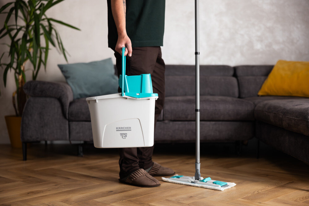 Osoba trzyma wiadro z logo KARCHER TTS, obok znajduje się mop płaski na drewnianej podłodze. W tle widoczna jest szara kanapa z poduszkami i roślina w doniczce.
