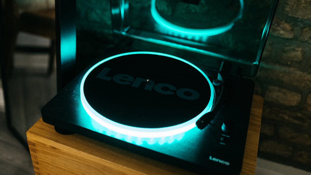 Czarny gramofon z podświetleniem LED, na którym widoczny jest napis \'Lenco\'. Urządzenie stoi na drewnianej powierzchni, a w tle widać ceglaną ścianę.