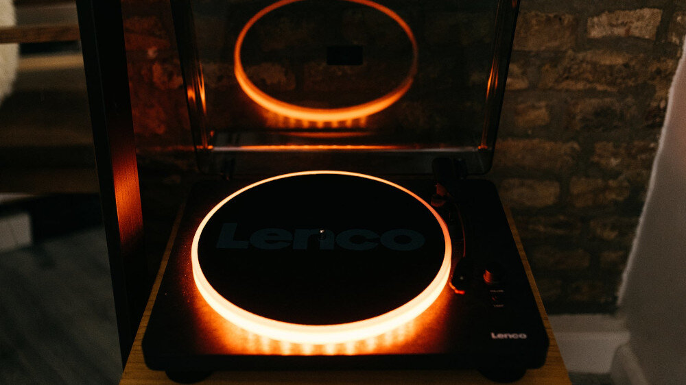 Gramofon z podświetleniem LED, na którym widoczne jest logo \'Lenco\'. Urządzenie stoi na drewnianej powierzchni, a w tle widać ceglaną ścianę.