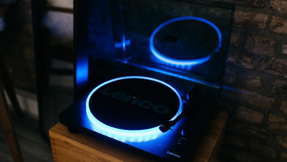 Gramofon z podświetleniem LED w kolorze niebieskim, z widocznym logo \'Lenco\' na talerzu. Urządzenie stoi na drewnianej powierzchni, a w tle widać ceglaną ścianę.