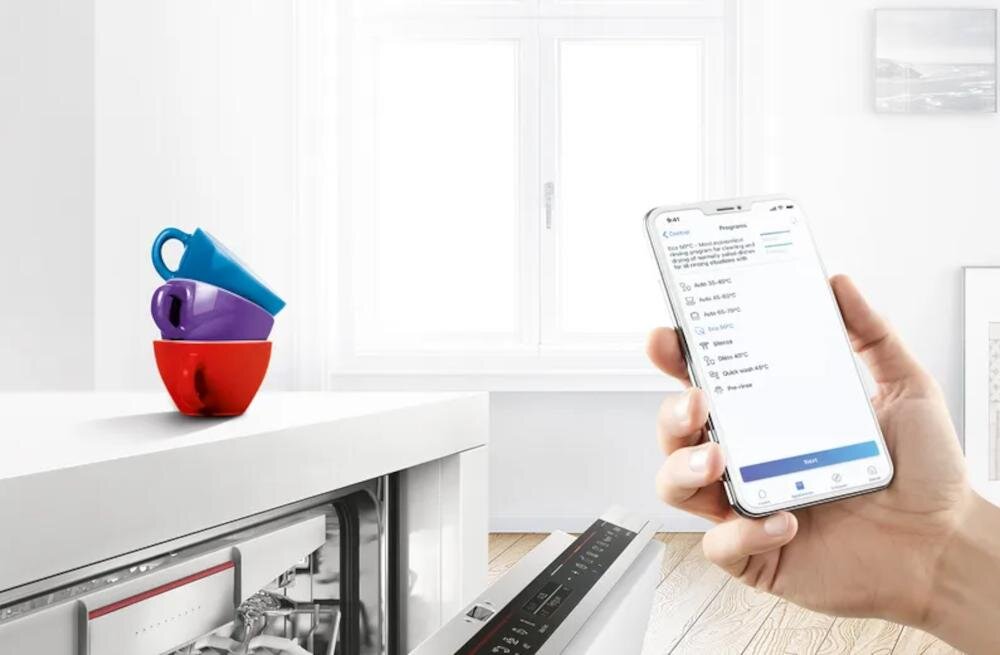 Na obrazie widać otwartą zmywarkę z widocznym panelem sterowania oraz rękę trzymającą smartfon z aplikacją. Na blacie stoją trzy kolorowe kubki ułożone jeden na drugim.