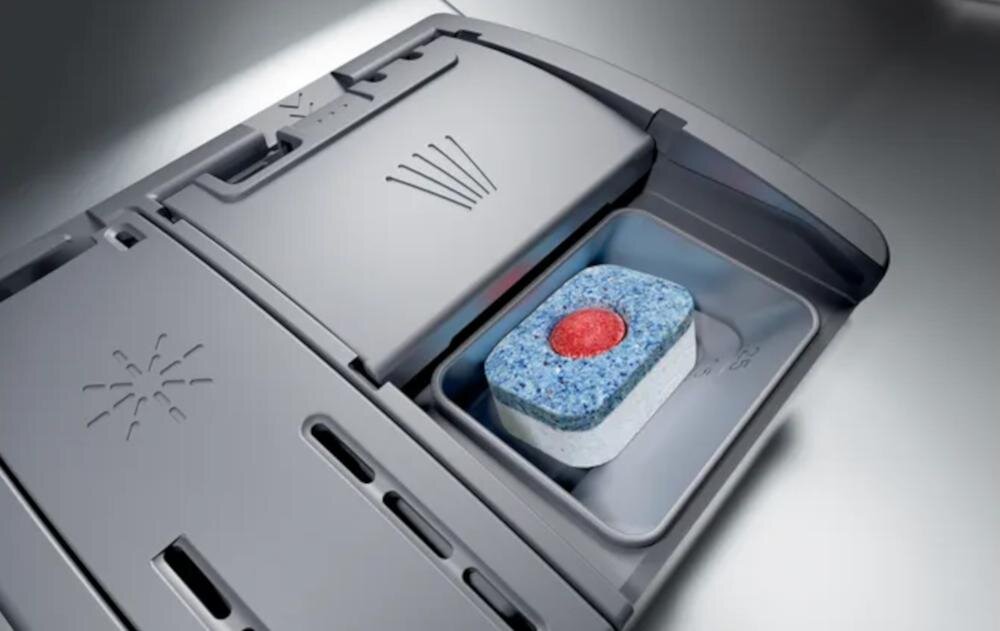 Zdjęcie przedstawia otwarty pojemnik na detergent w zmywarce, w którym znajduje się niebieska tabletka z czerwonym środkiem. Obok pojemnika widoczne są symbole wskazujące na funkcje urządzenia.