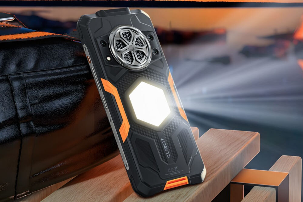 Smartfon CUBOT KING KONG Power 5 16/128GB 6.88 120Hz Czarny Zbliżenie na tylną część pancernego smartfona Cubot z włączoną dużą, sześciokątną latarką. Smartfon leży na drewnianym blacie, a w tle widać ciemną torbę i źródło światła