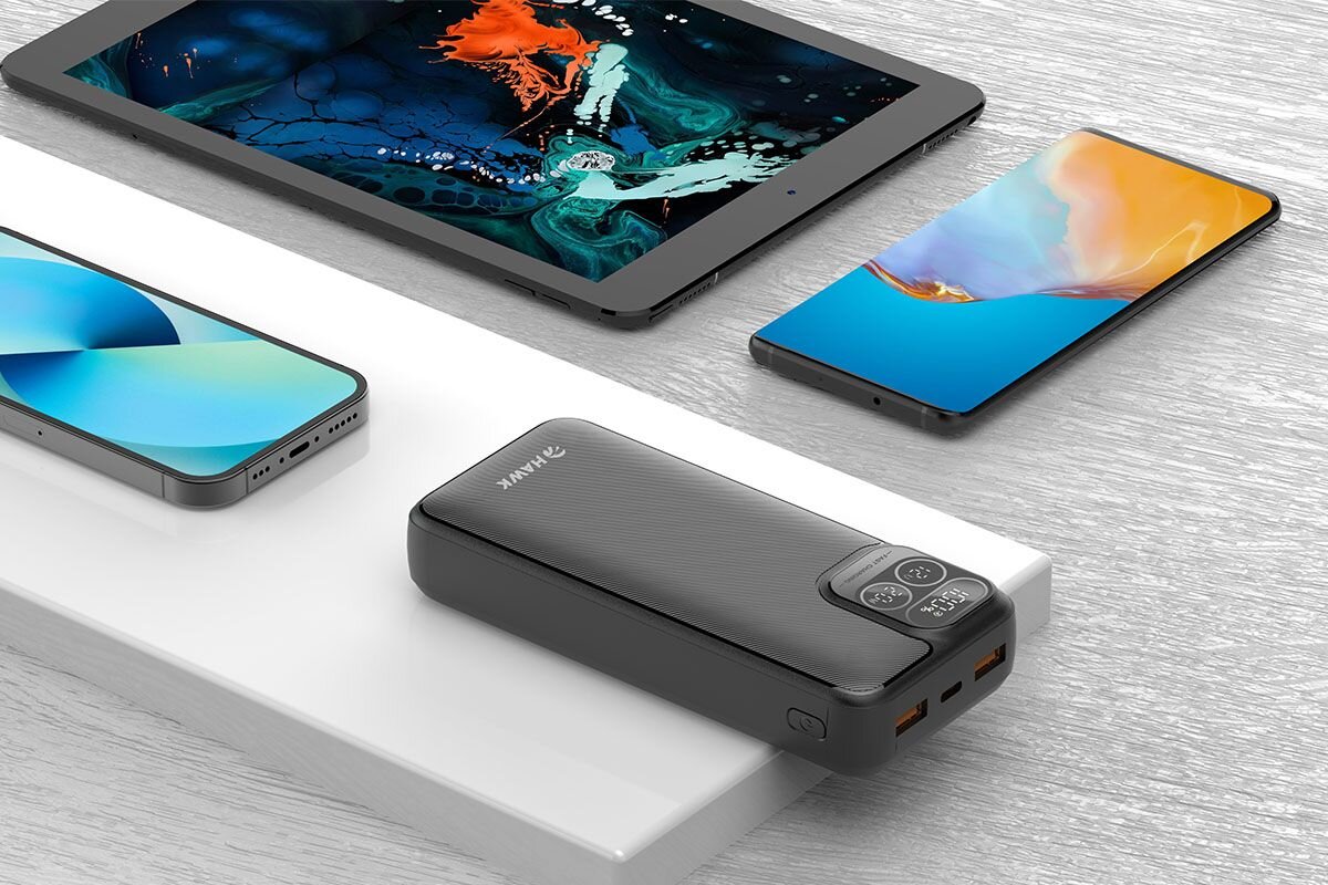 Na obrazie widoczny jest czarny powerbank z logo \'HAWK\', umieszczony na białej powierzchni, obok trzech urządzeń elektronicznych: tabletu i dwóch smartfonów. Powerbank ma wyświetlacz pokazujący stan naładowania oraz kilka portów do ładowania.