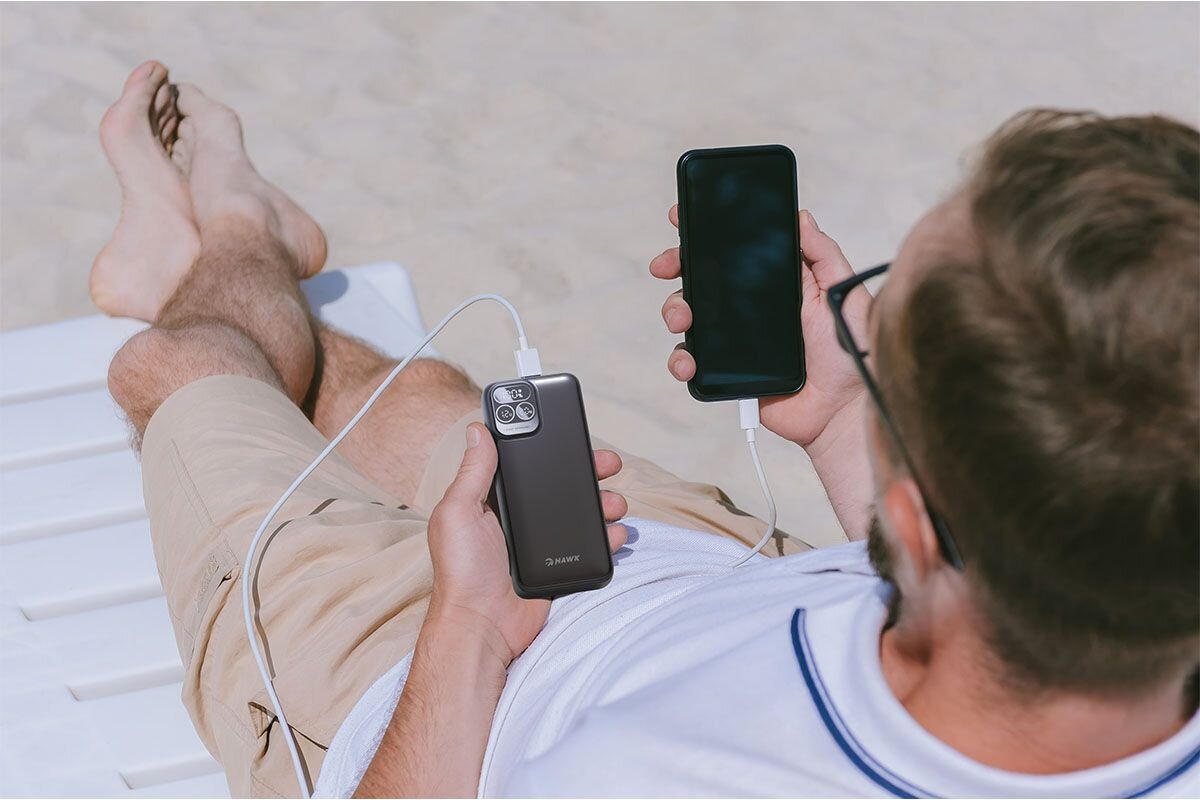 Osoba leży na leżaku na plaży, trzymając w rękach smartfon i czarny powerbank z logo \'HAWK\'. Powerbank jest podłączony do smartfona za pomocą kabla.