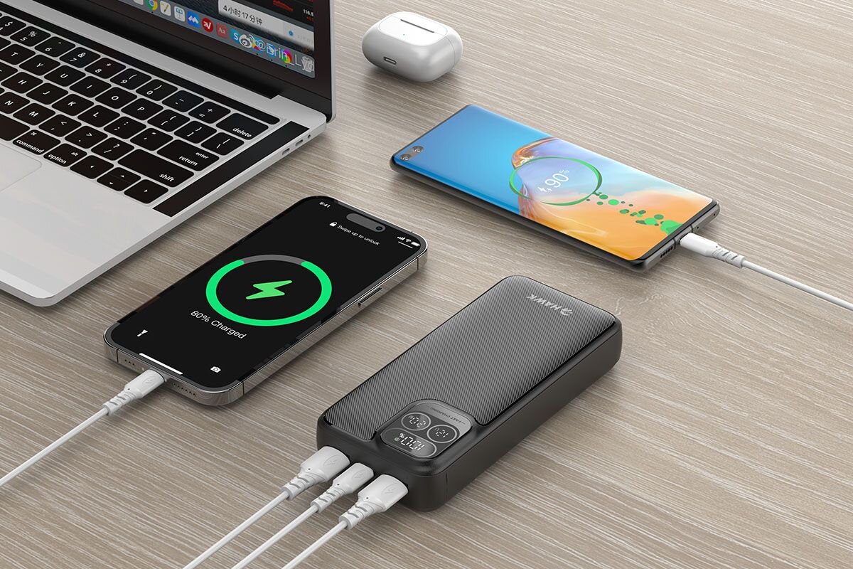 Na drewnianym stole znajduje się czarny powerbank z logo \'HAWK\', do którego podłączone są dwa smartfony i laptop. Na ekranie jednego z telefonów widoczny jest napis \'80% Charged\'.