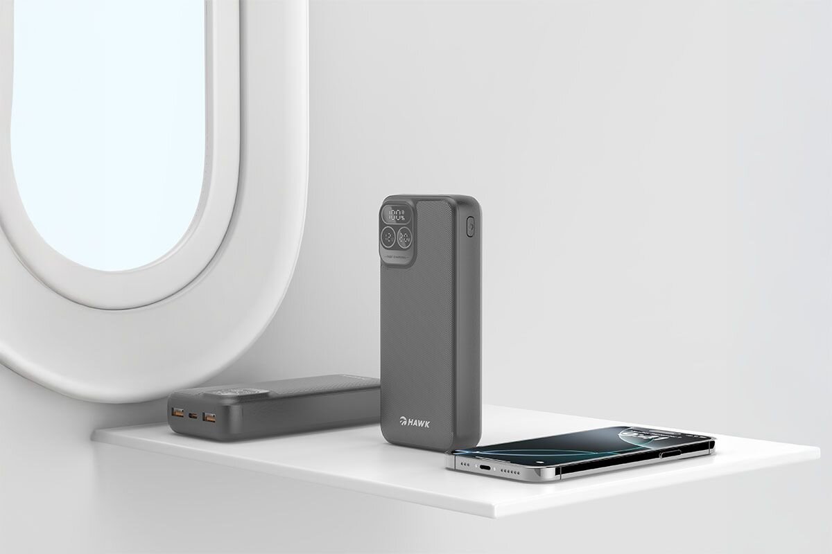 Na białej półce znajduje się czarny powerbank z logo \'HAWK\', obok którego leży smartfon. W tle widoczne jest okno samolotu.