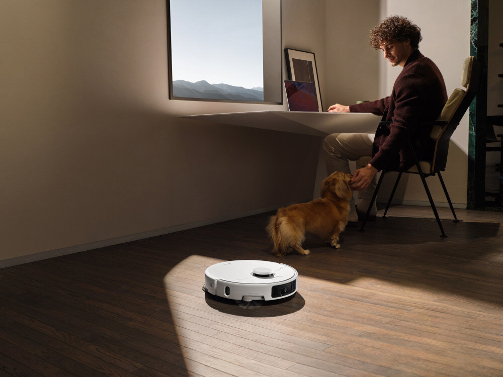 W pomieszczeniu z drewnianą podłogą znajduje się robot sprzątający oraz osoba siedząca przy biurku z psem. Przez okno widać górski krajobraz.