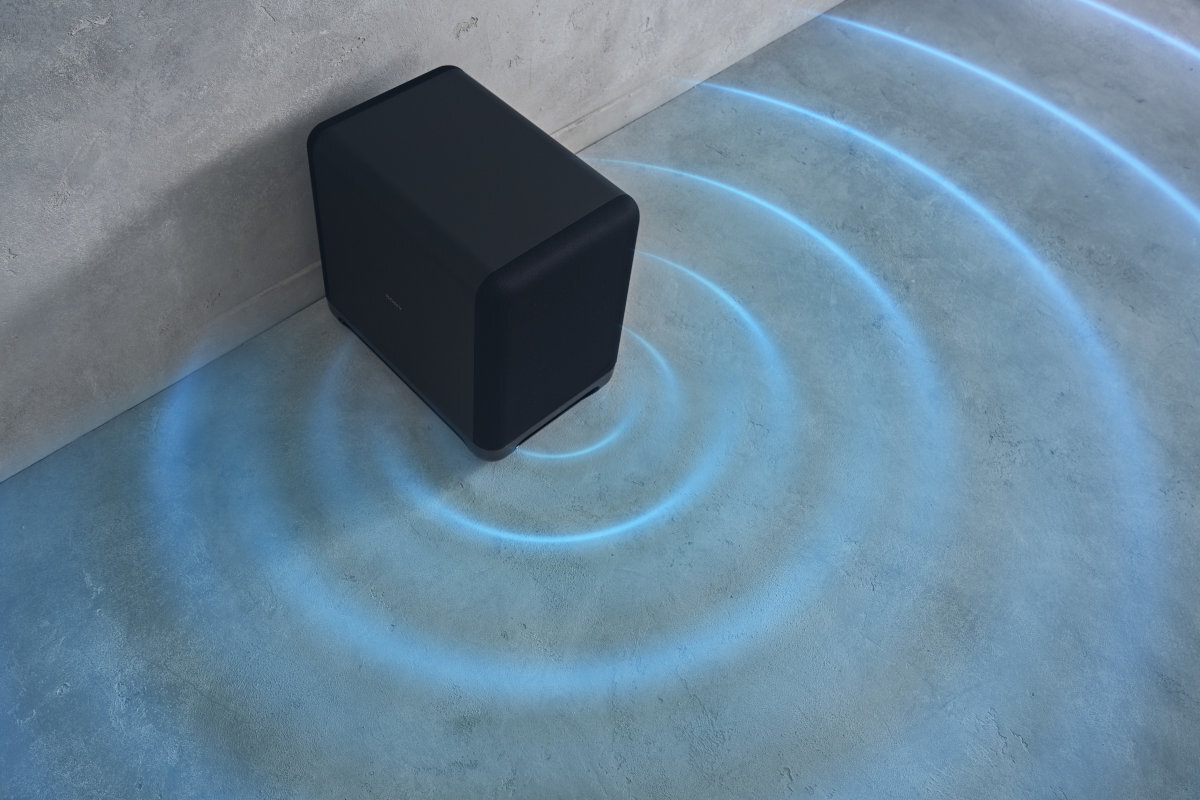 Czarny subwoofer stoi na podłodze przy ścianie, emitując niebieskie fale dźwiękowe widoczne na podłodze. Na urządzeniu widoczne jest logo producenta.