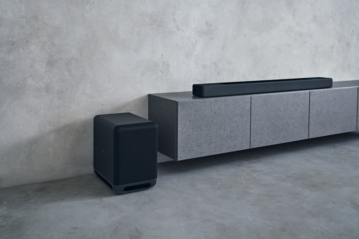 Na obrazie widoczny jest czarny subwoofer obok szarej szafki z soundbarem na górze. Tło stanowi jasna, betonowa ściana.