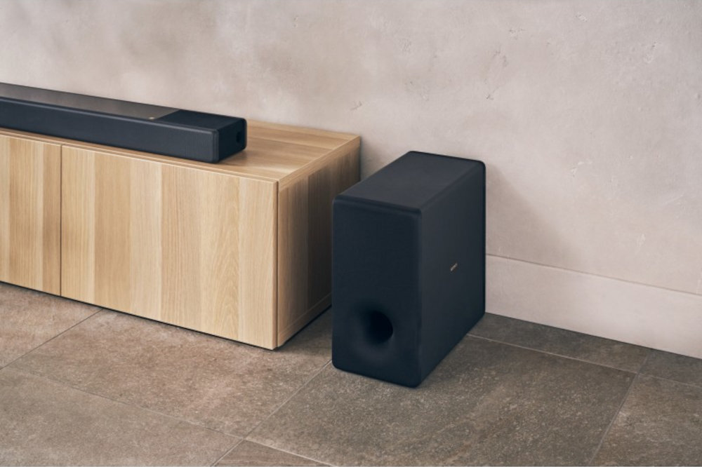 Na obrazie widać czarny subwoofer ustawiony obok drewnianej szafki, na której znajduje się soundbar. Subwoofer ma prostokątny kształt i widoczne logo producenta.