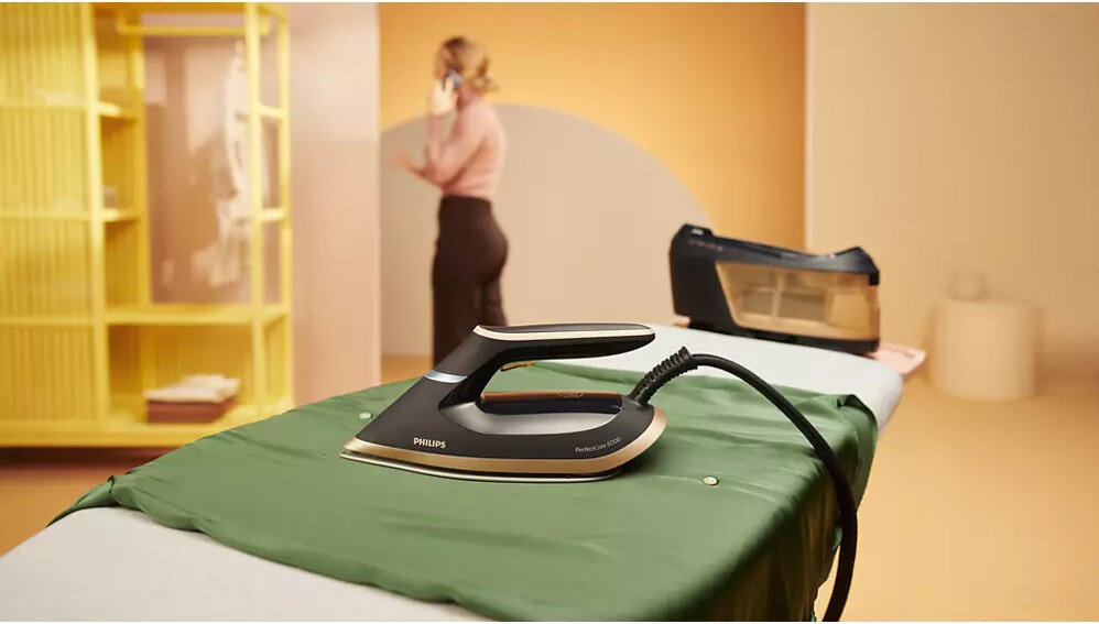 Na desce do prasowania znajduje się żelazko z napisem \'PHILIPS PerfectCare\'. W tle widoczna jest osoba rozmawiająca przez telefon oraz stacja parowa.