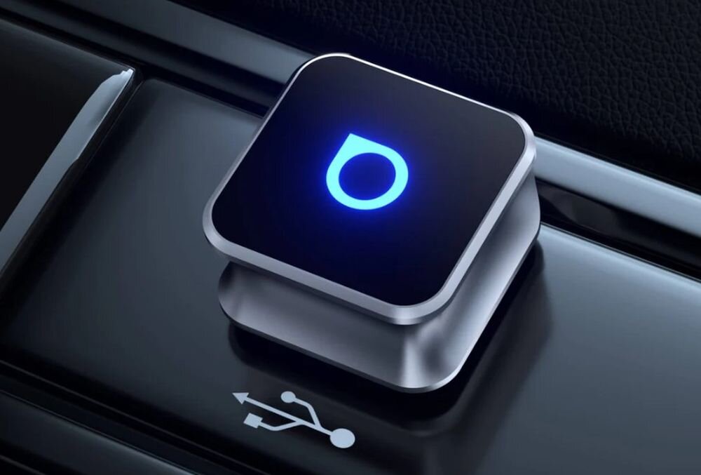 Kwadratowe urządzenie z podświetlonym niebieskim symbolem na czarnym tle, umieszczone na błyszczącej powierzchni. Obok widoczny jest symbol USB.
