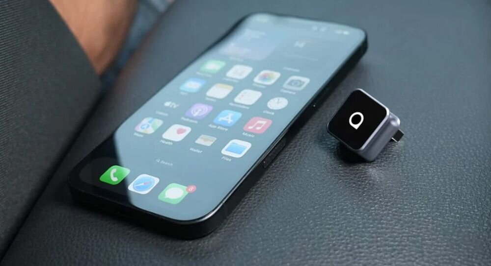Na zdjęciu widać smartfon z wyświetlonymi ikonami aplikacji oraz małe, czarne urządzenie z białym symbolem na górze, leżące na ciemnej powierzchni.
