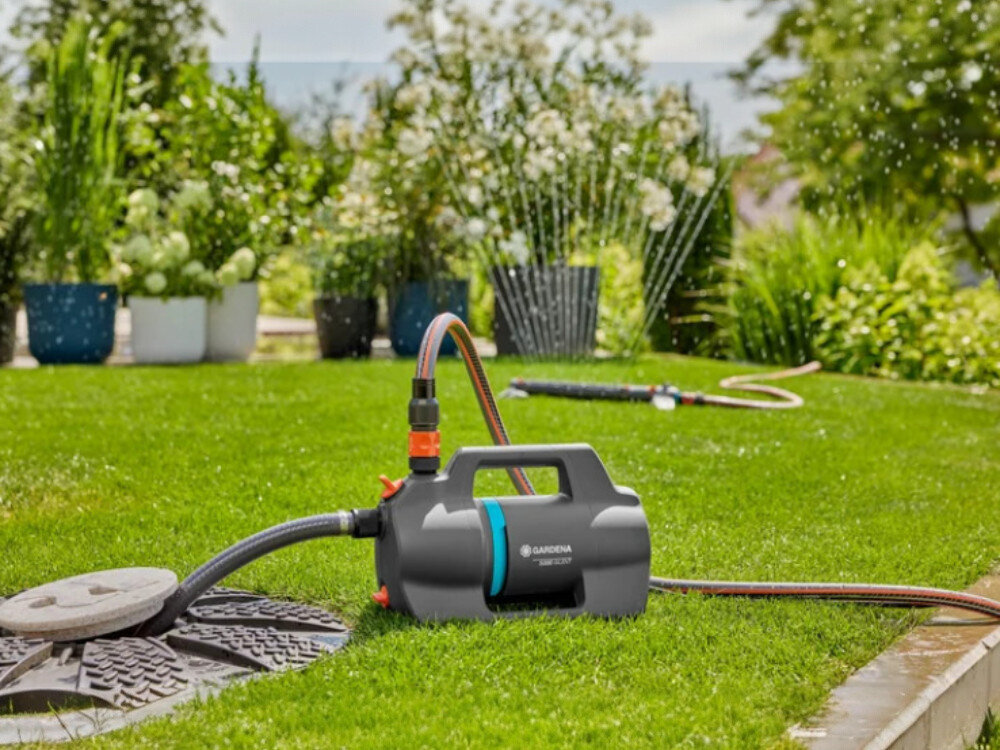 Na trawniku znajduje się elektryczna pompa do wody z widocznym logo GARDENA, podłączona do węża ogrodowego. W tle widać rośliny i działający zraszacz.