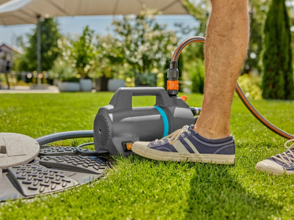 Na trawniku stoi elektryczna pompa do wody z podłączonym wężem ogrodowym. Obok pompy widoczne są nogi osoby w sportowych butach.