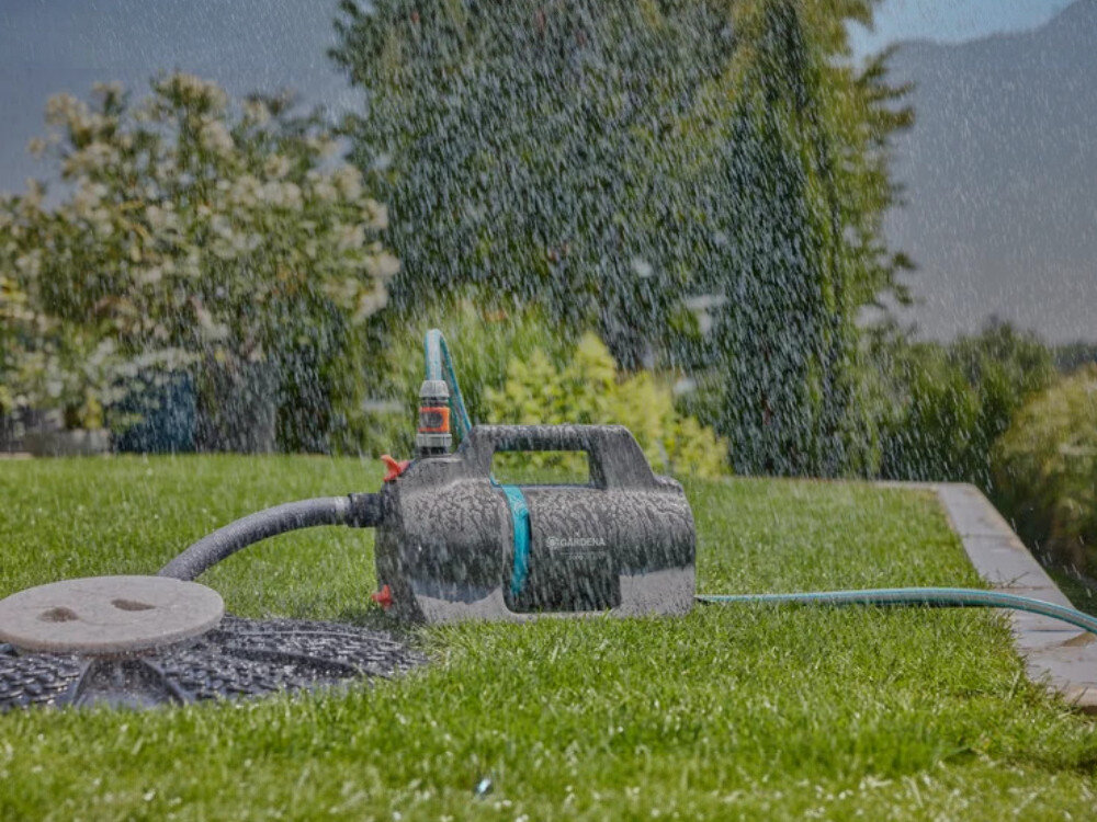 Na trawniku znajduje się elektryczna pompa do wody z widocznym logo \'GARDENA\', podłączona do węża ogrodowego. W tle widać roślinność i padający deszcz.