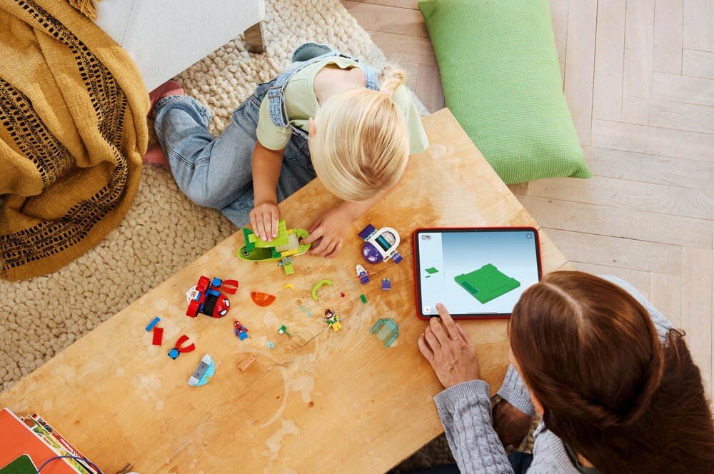 Dwoje dzieci bawi się zestawem klocków LEGO na drewnianym stole, obok leży tablet z wyświetlonym modelem 3D. Na stole znajdują się różnokolorowe elementy LEGO oraz małe figurki.