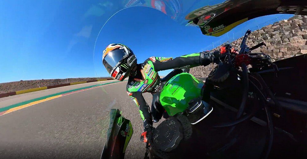 Osoba w kombinezonie motocyklowym jedzie na zielonym motocyklu po torze wyścigowym. Obraz jest zniekształcony, co sugeruje użycie kamery sferycznej.