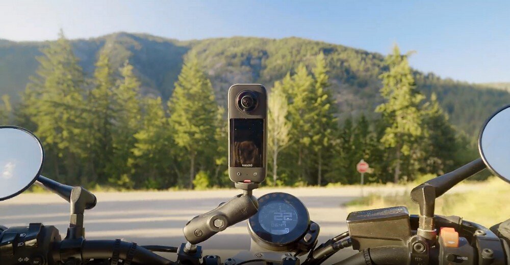 Kamera sferyczna zamontowana na motocyklu, w tle widoczne są drzewa i góry. Na ekranie kamery odbija się krajobraz.