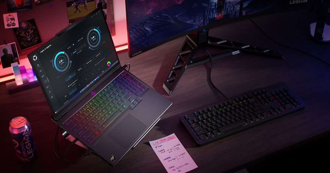 Na biurku znajduje się laptop z podświetlaną klawiaturą, obok którego leży klawiatura zewnętrzna i monitor. Na biurku widoczna jest także puszka z napisem \'Legion\' oraz kartka z notatkami.