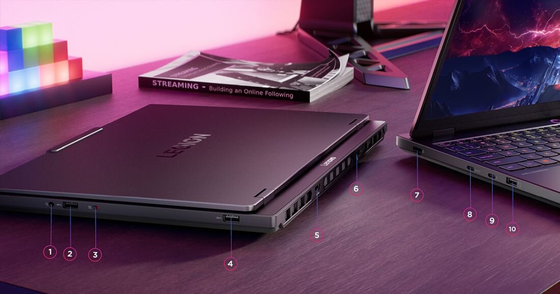 Na biurku znajdują się dwa laptopy z widocznymi portami i logo \'LEGION\' na jednym z nich. Obok leży książka z napisem \'STREAMING - Building an Online Following\'.