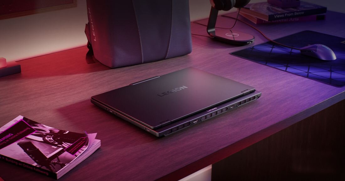 Na biurku znajduje się zamknięty laptop z widocznym logo \'LEGION\'. Obok laptopa leży książka i mysz komputerowa.
