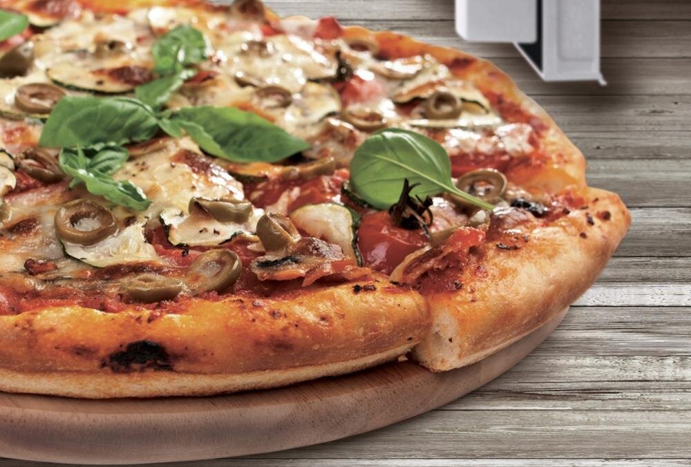 Na drewnianej desce znajduje się pizza z oliwkami, plasterkami pomidora i liśćmi bazylii. W tle widoczna jest część kuchenki mikrofalowej.