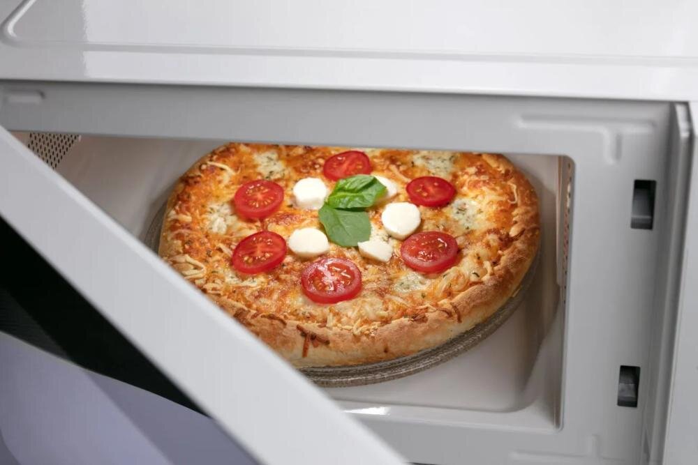 Otwarta kuchenka mikrofalowa z widoczną pizzą na talerzu obrotowym. Pizza jest udekorowana plasterkami pomidora, mozzarellą i liśćmi bazylii.