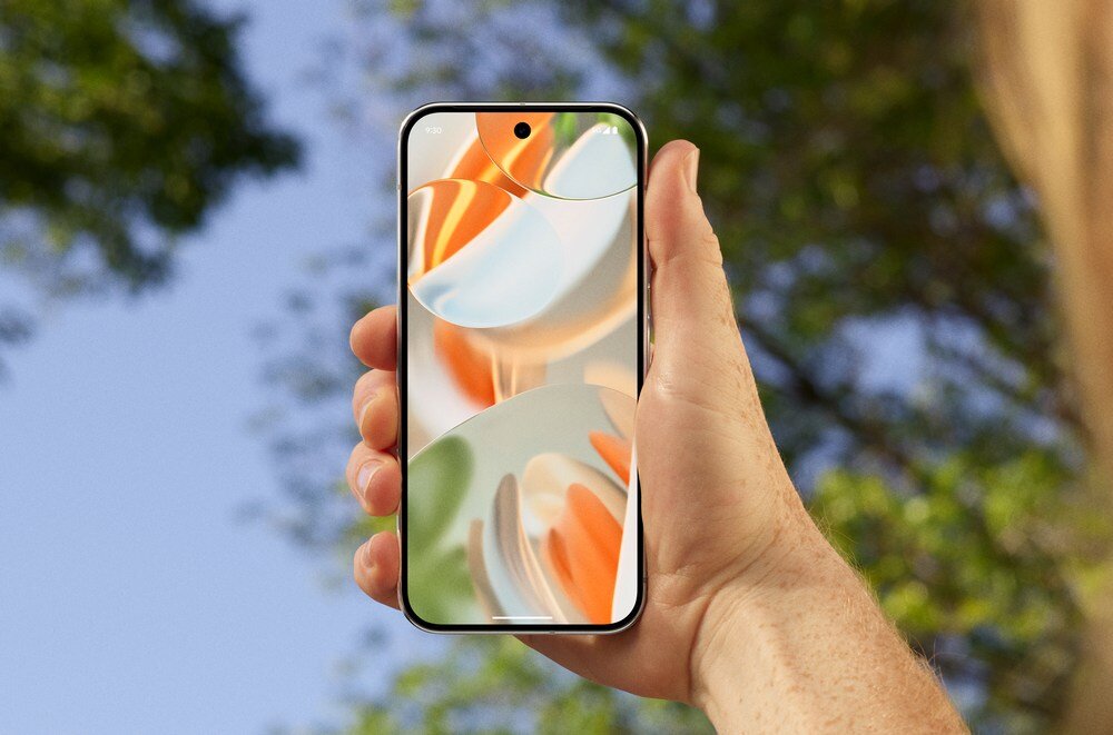 Osoba trzyma smartfon z kolorowym, abstrakcyjnym wzorem na ekranie. W tle widać rozmyte drzewa i niebo.