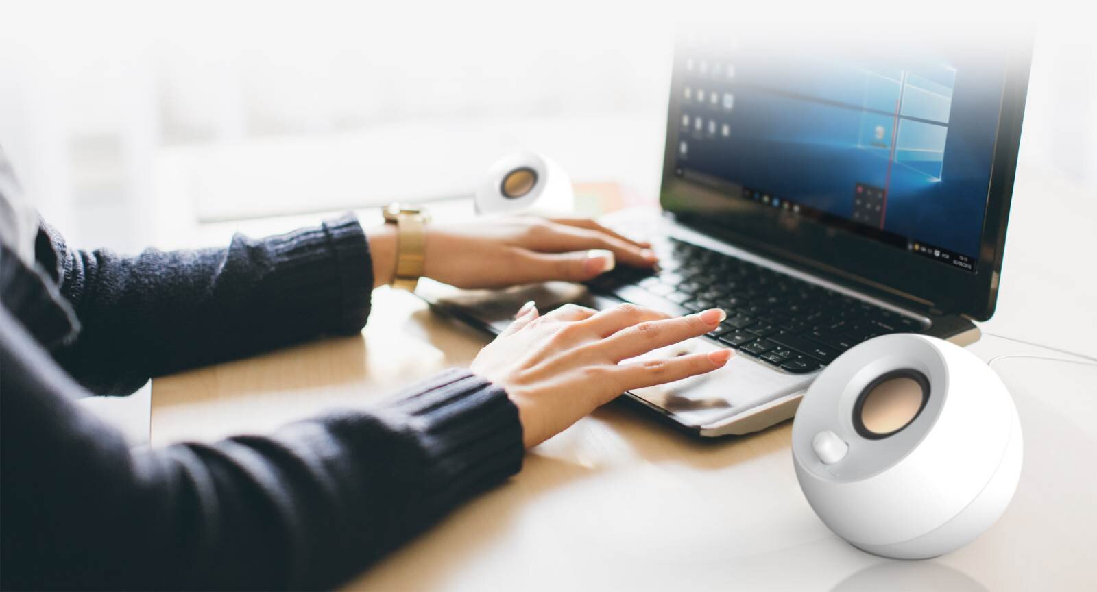 Na biurku stoi otwarty laptop z widocznym ekranem, obok którego znajdują się białe głośniki o kulistym kształcie. Osoba siedząca przy biurku używa klawiatury laptopa.