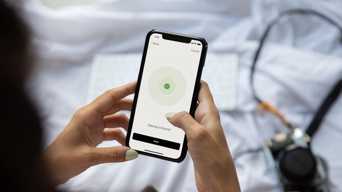 Osoba trzyma smartfon z aplikacją pokazującą komunikat \'Device is found\'. W tle widoczna jest klawiatura i aparat fotograficzny.