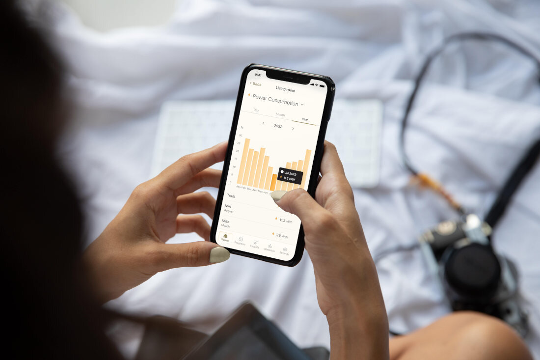 Osoba trzyma smartfon z aplikacją pokazującą wykres zużycia energii z napisem \'Power Consumption\'. W tle widoczna jest klawiatura i aparat fotograficzny.
