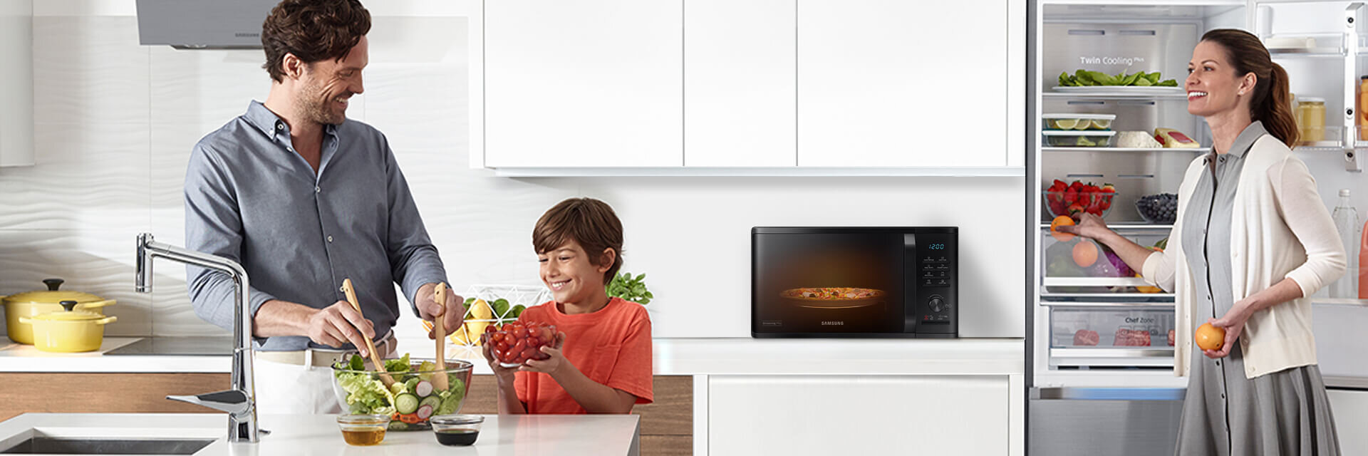 W kuchni znajduje się kuchenka mikrofalowa z widocznym logo \'Samsung\' na blacie. Obok niej stoi otwarta lodówka z różnymi produktami spożywczymi.