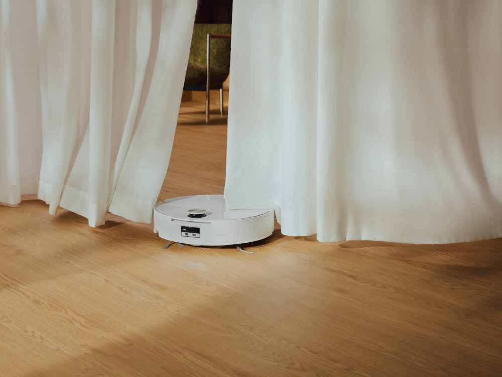 Biały robot sprzątający porusza się po drewnianej podłodze, częściowo zasłonięty białymi zasłonami. W tle widoczne są nogi krzesła.