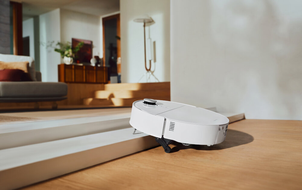 Na drewnianej podłodze znajduje się biały robot sprzątający, który pokonuje niewielki próg. W tle widoczny jest nowoczesny salon z sofą i lampą stojącą.