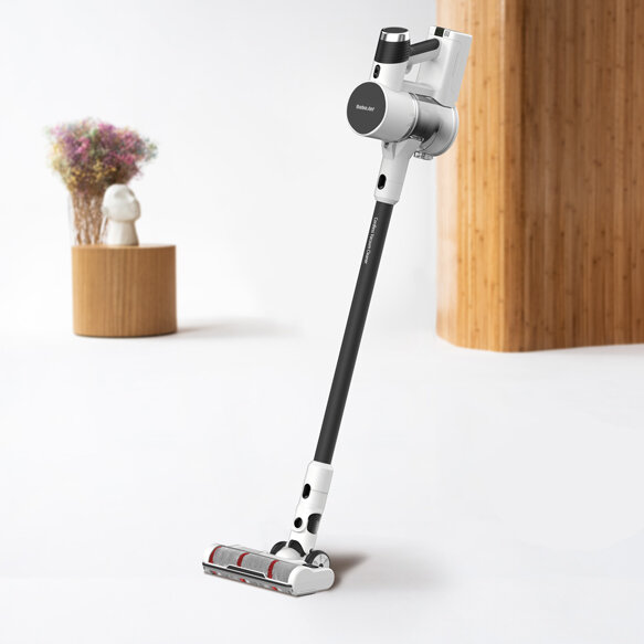 Na obrazie widoczny jest odkurzacz pionowy z napisem \'ROBOJET\' na korpusie, stojący na białej podłodze. W tle znajduje się drewniany element dekoracyjny oraz wazon z kwiatami.