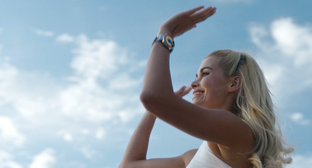 Osoba z długimi blond włosami nosi smartwatch na nadgarstku, patrząc w górę na tle nieba z chmurami.